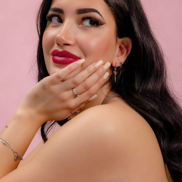 Mujer con cabello largo y oscuro usando joyas plateadas con cristales rojos sobre un fondo rosa