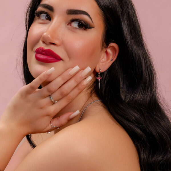 Mujer con cabello largo y negro, labios rojos y pendiente plateado con cristal rojo frente a fondo rosa