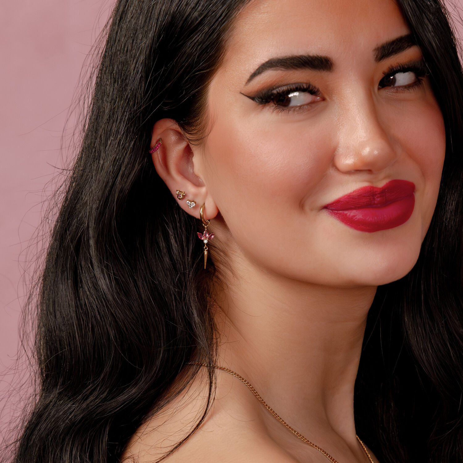 Mujer con cabello negro usando varios pendientes, incluido un aro botánico de rubí sobre fondo rosa.