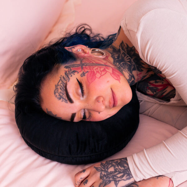 Persona con tatuajes recostada en un cojín para piercings negro sobre ropa de cama rosa