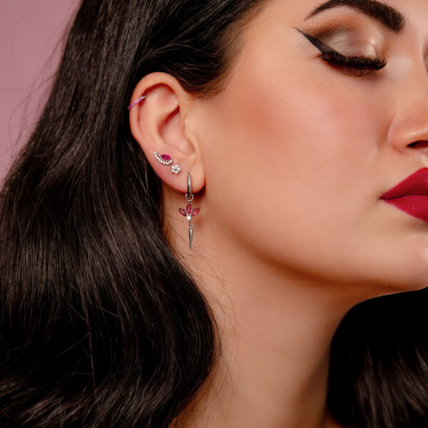 Primer plano de una oreja con varios piercings, incluyendo un clicker plateado con cristales rojos, sobre fondo rosa.