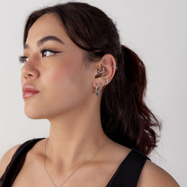 Mujer de cabello oscuro lleva un septum clicker negro y varios pendientes de diferentes formas sobre un fondo blanco.
