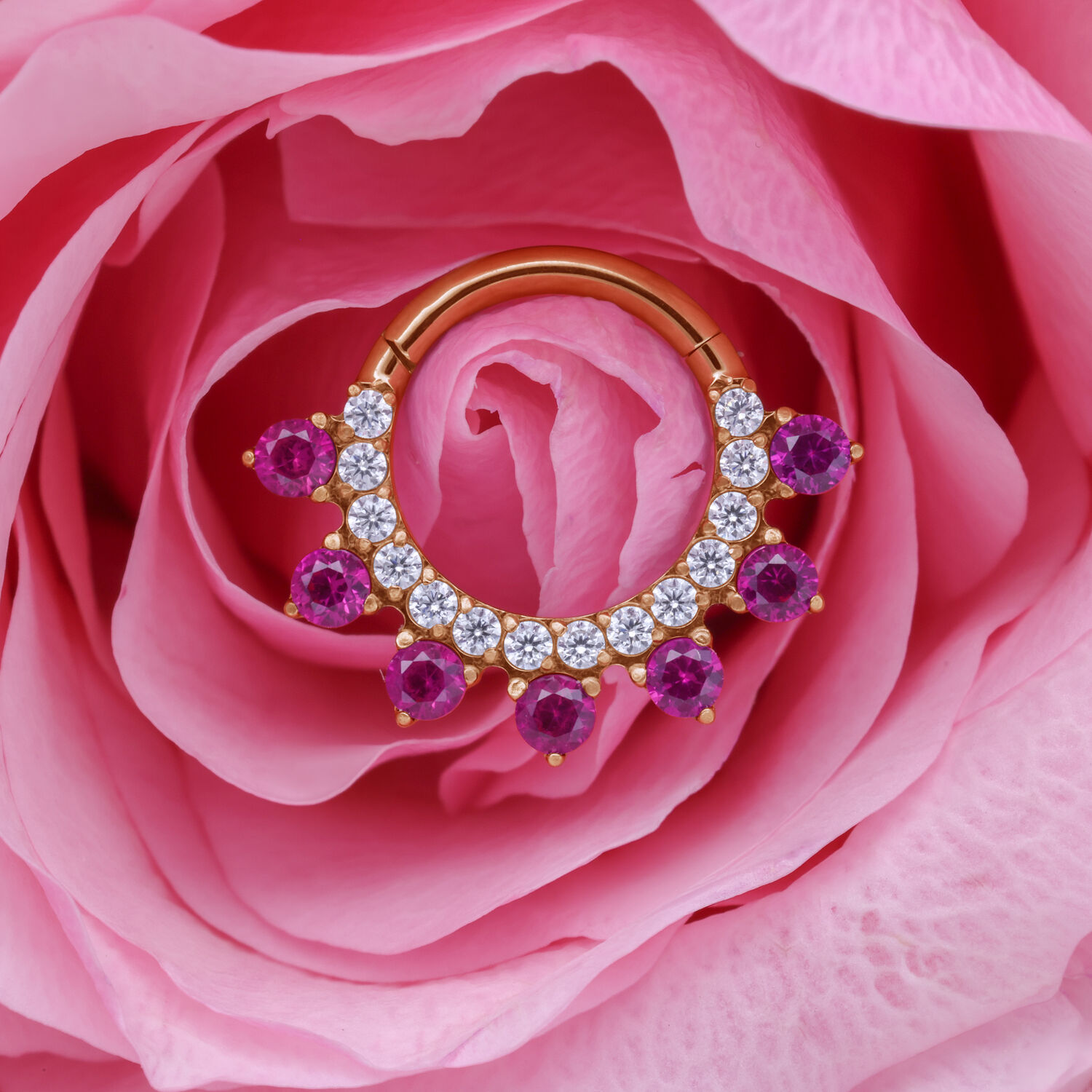 Anillo de piercing dorado con cristales rojos y claros, colocado en una flor rosa.