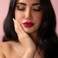 Mujer con cabello negro y piercing nasal plateado con cristal rojo frente a fondo rosa