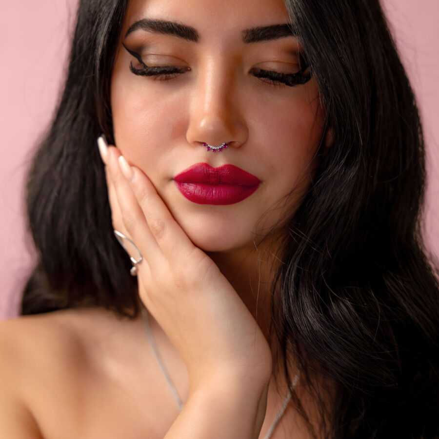 Mujer con cabello negro y piercing nasal plateado con cristal rojo frente a fondo rosa