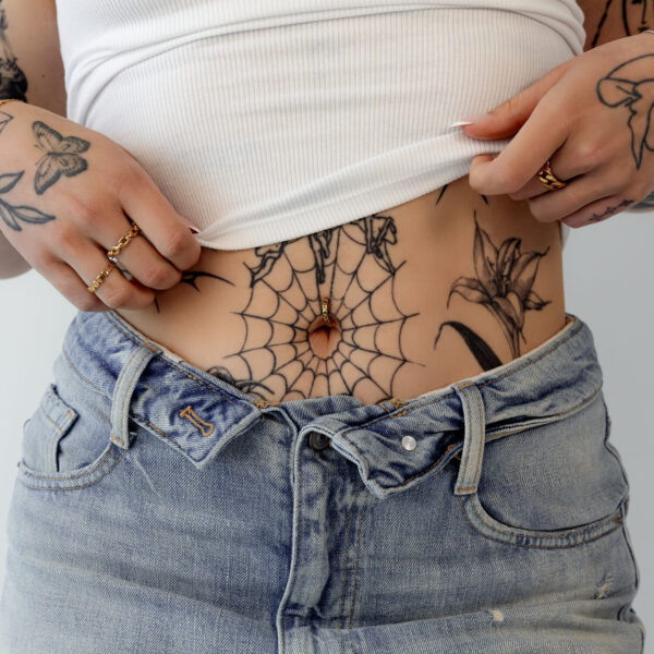 Piercing de ombligo con estampado de leopardo dorado, usado por persona con tatuajes y jeans.