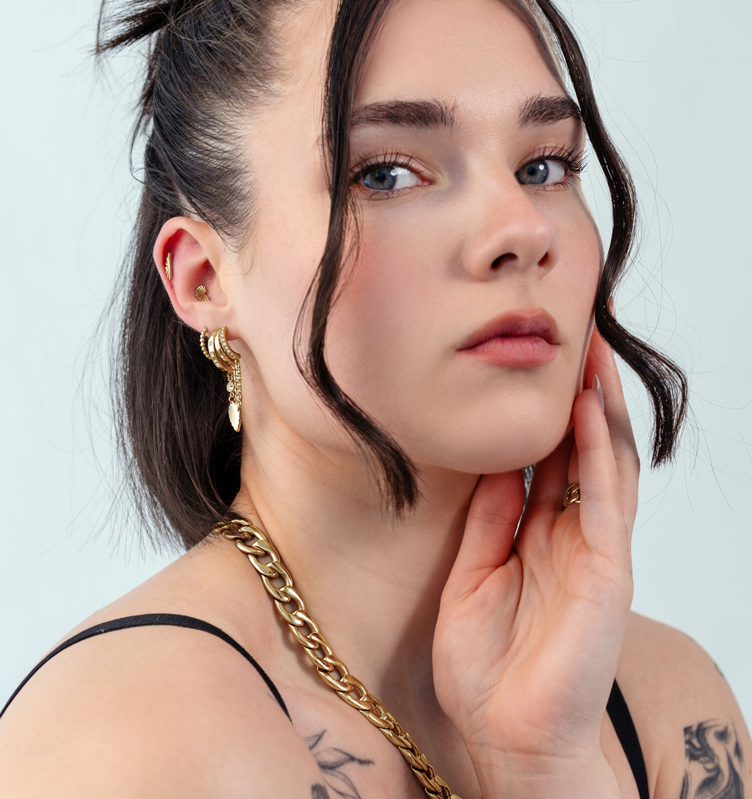 Mujer con pendientes dorados en forma de hoja, lleva un collar dorado, sobre un fondo claro.