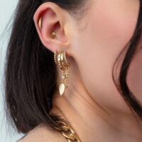 Primer plano de una oreja con pendientes de hoja Riffle dorados y colgante de cadena, cabello oscuro de fondo