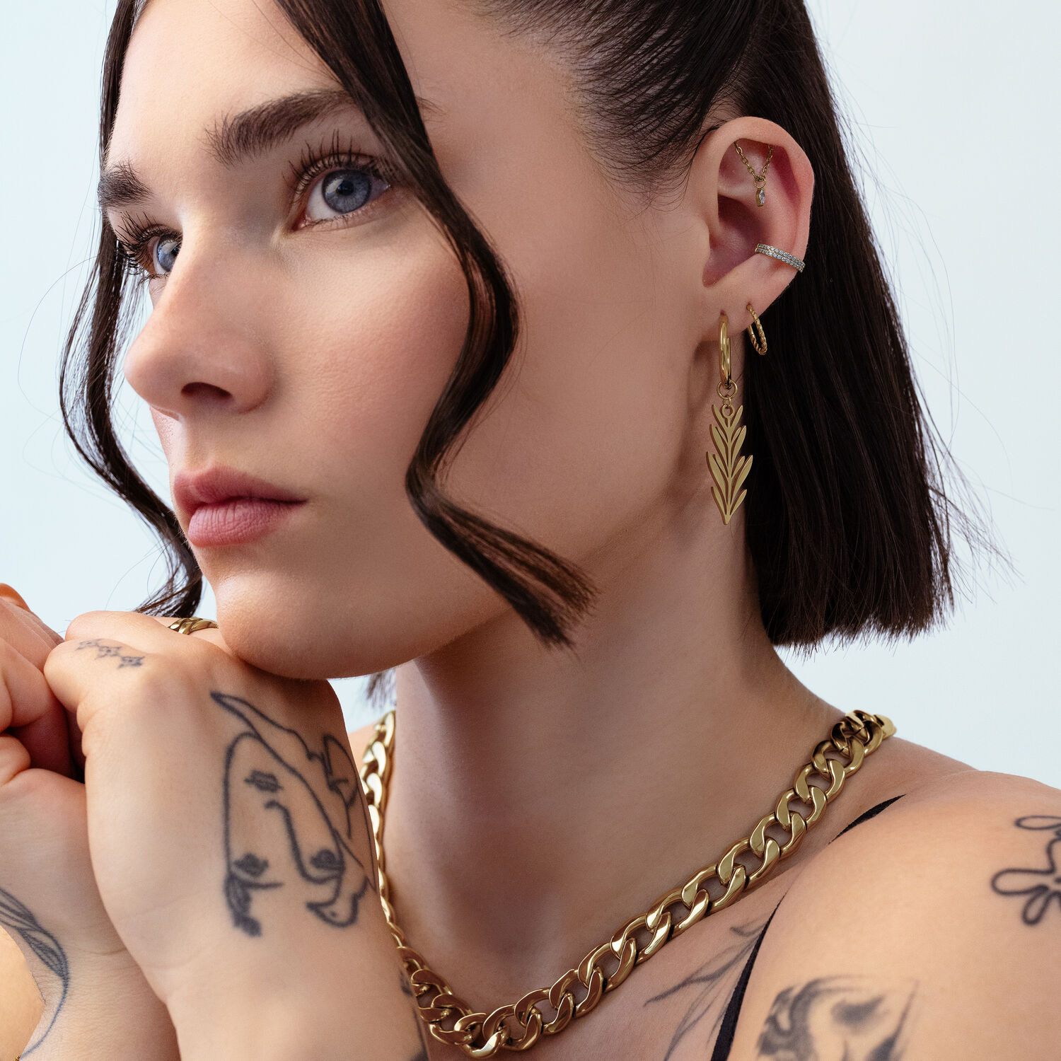 Aros Lovely Branch dorados en la oreja Mujer con aros dorados y joyas de cadena frente a un fondo claro