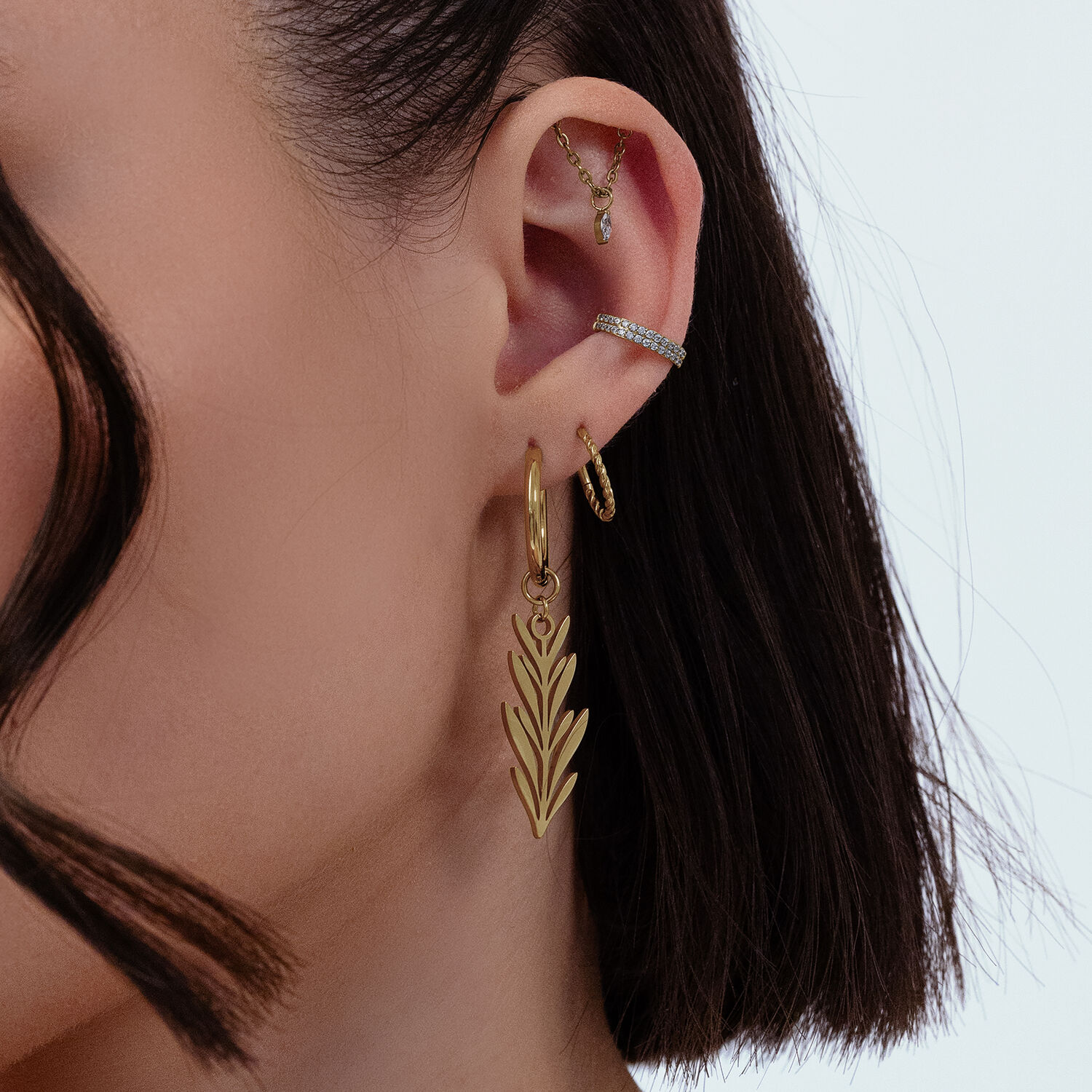 Pendientes dorados con diseño de hoja Primer plano de una oreja con varios pendientes dorados, incluyendo un gran diseño de hoja, sobre fondo blanco