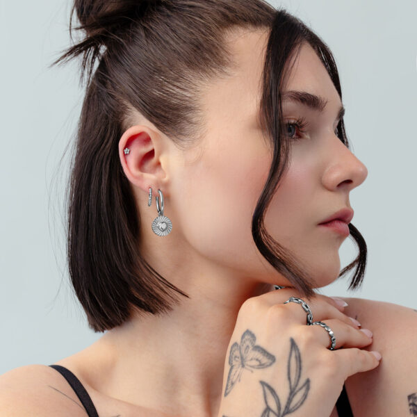 Mujer con cabello corto y oscuro lleva aros plateados con diseño de corazón y tatuajes sobre fondo gris claro.