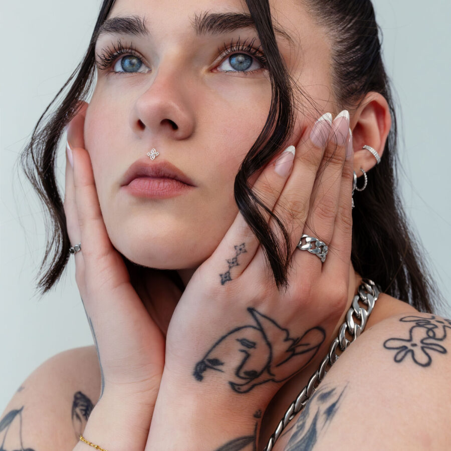 Mujer con manos tatuadas tocando su rostro, lleva un adorno de flor plateado en el piercing de la nariz