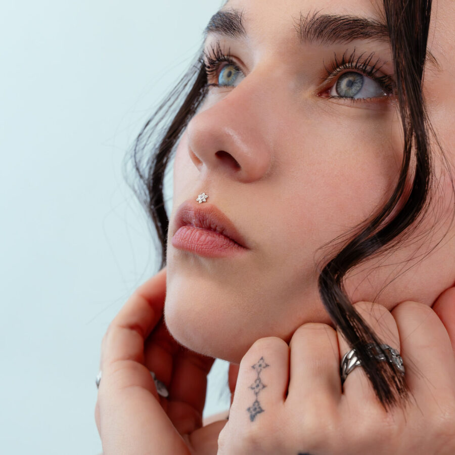 Primer plano de un rostro con un piercing de flor plateada en la nariz, fondo azul claro