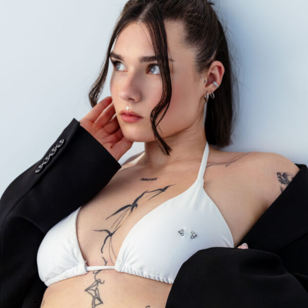Mujer con top de bikini blanco y blazer negro, tatuajes visibles, sobre fondo claro