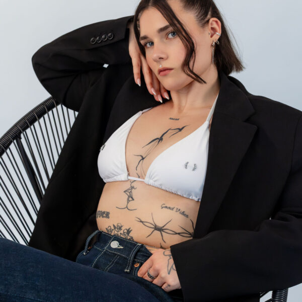 Mujer con top de bikini blanco y blazer negro en una silla, con el torso tatuado