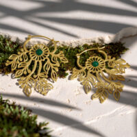 Pendientes dorados con diseño de hojas Dos pendientes dorados con diseño de hojas y centro verde sobre fondo claro, rodeados de musgo