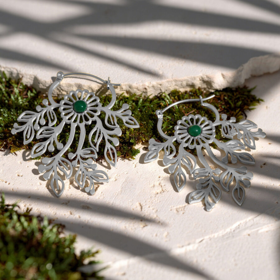 Dos pendientes plateados con ornamentos delicados y piedras verdes sobre una superficie blanca con musgo