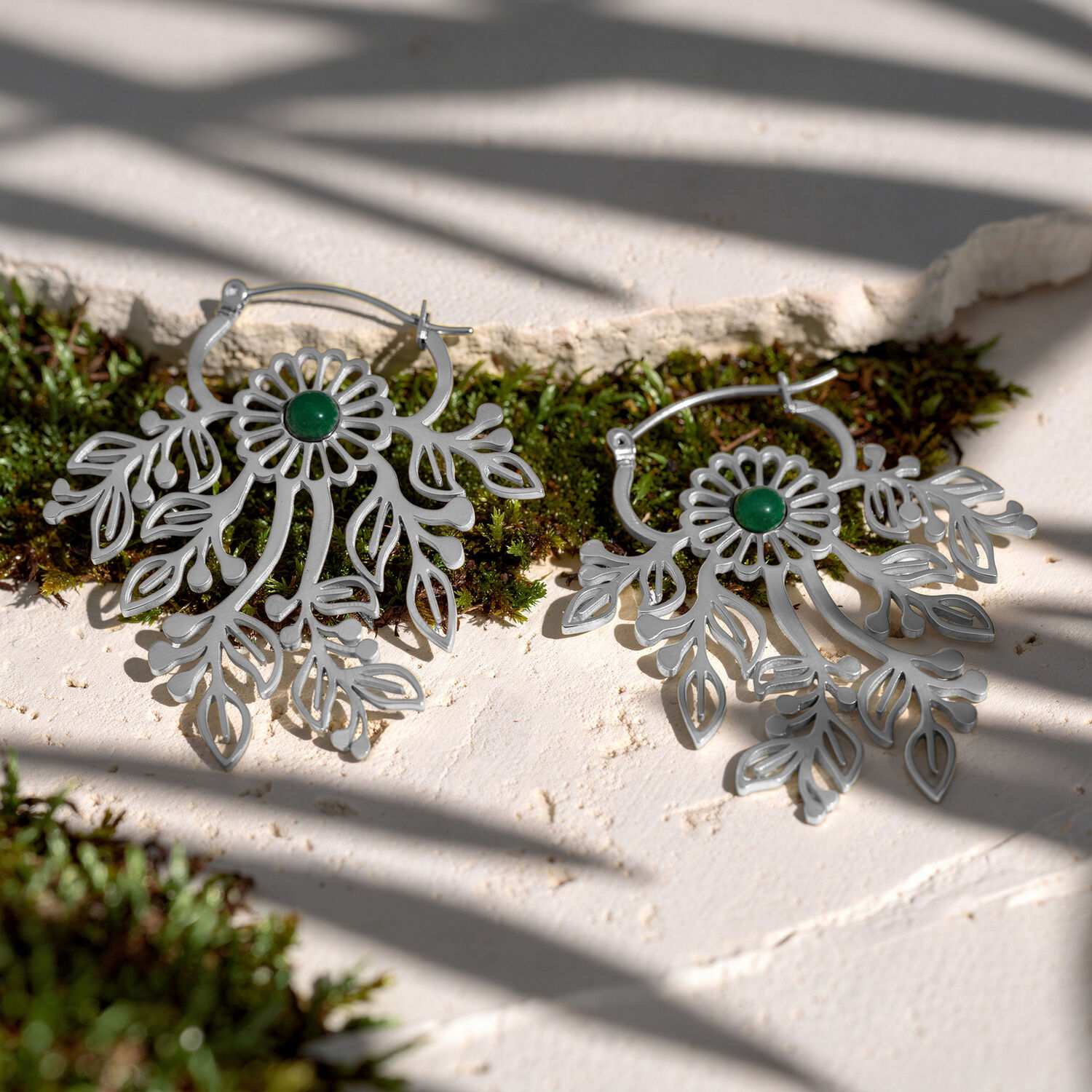 Pendientes de ornamentos plateados con piedras verdes Dos pendientes plateados con ornamentos delicados y piedras verdes sobre una superficie blanca con musgo
