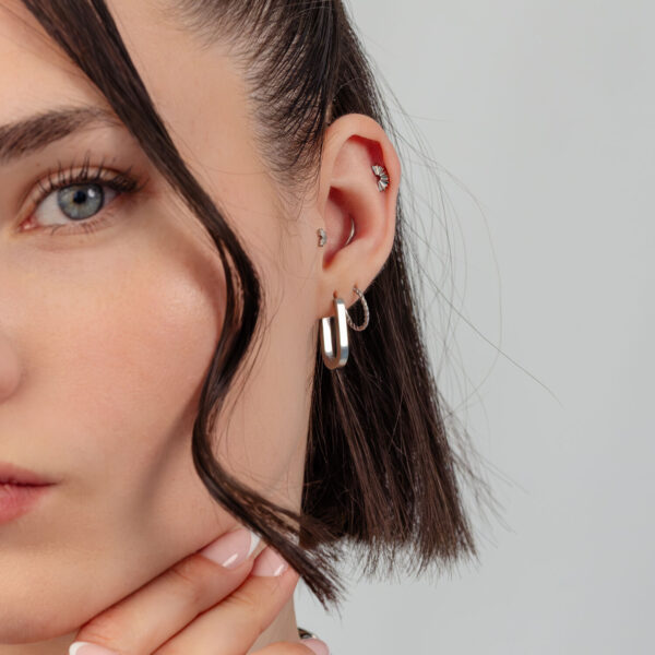 Primer plano de una oreja con aros de plata y varios piercings delante de un fondo gris.