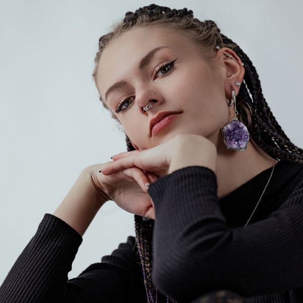 Joven con trenzas, pendiente de amatista, piercing en la nariz, suéter negro frente a fondo gris