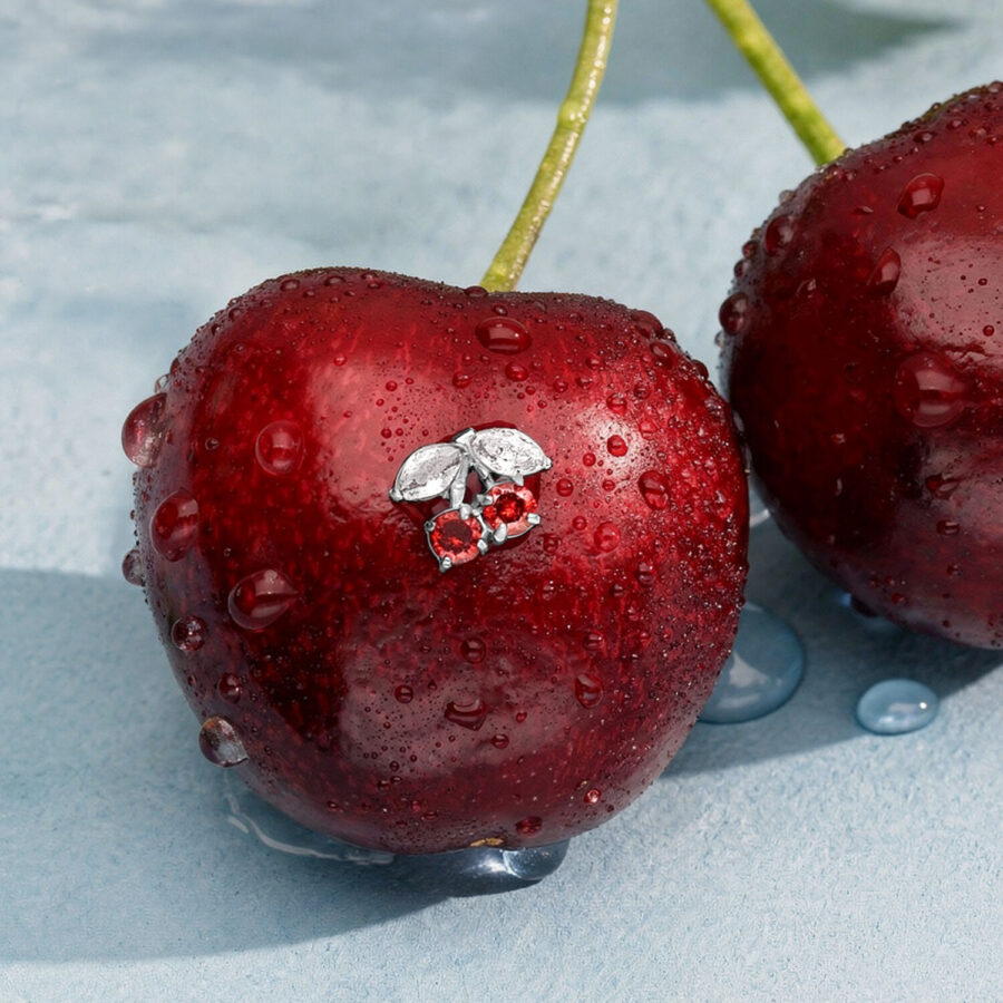 Colgante de cereza plateado con circonitas rojas sobre una cereza fresca, cubierto de gotas de agua.