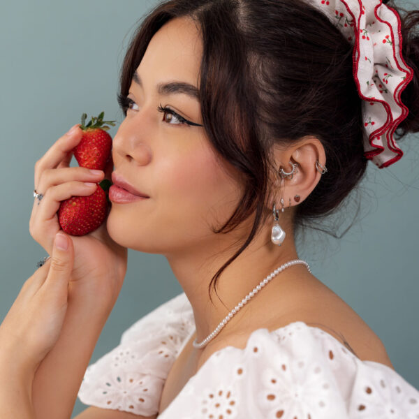 Mujer con vestido de encaje blanco, fresa en mano y pendientes de joyas frente a fondo azul