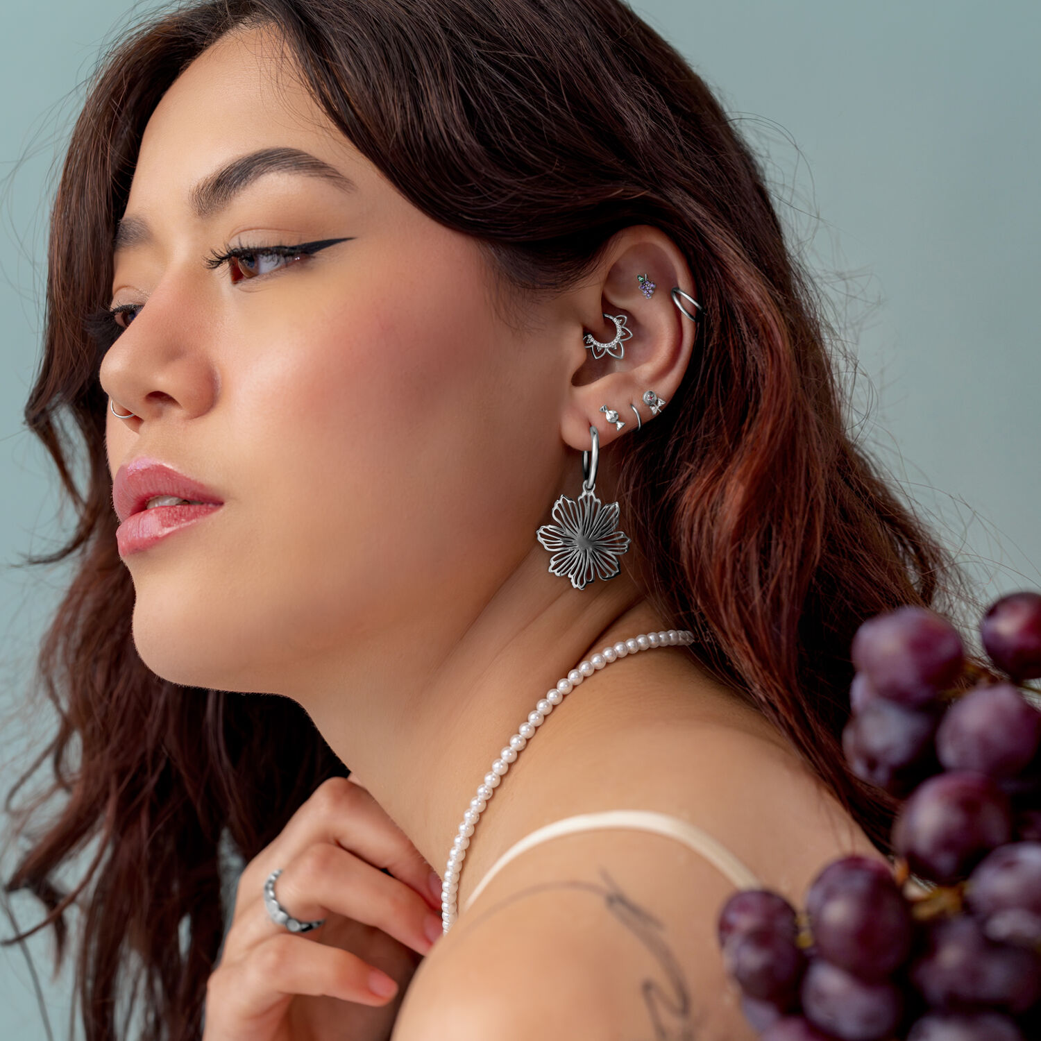 Ajuste a presión de uva plateada Mujer con varios piercings plateados en la oreja, lleva un collar de perlas y sostiene uvas.