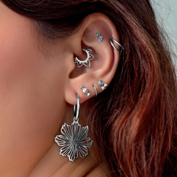 Primer plano de una oreja con varios piercings plateados, incluyendo un gran colgante de flor, sobre un fondo claro