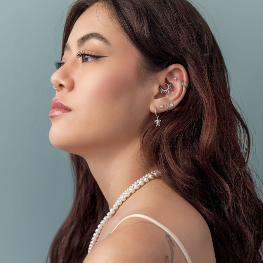 Mujer con cabello castaño lleva varios piercings plateados y un colgante de perlas frente a un fondo azul