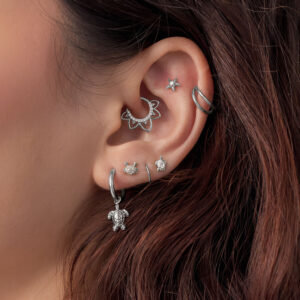 Primer plano de una oreja con varios accesorios de piercing plateados, incluyendo un diseño de tortuga, sobre cabello castaño.
