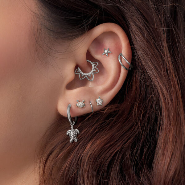 Primer plano de una oreja con varios accesorios de piercing plateados, incluyendo un diseño de tortuga, sobre cabello castaño.