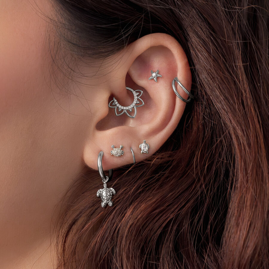 Primer plano de una oreja con varios accesorios de piercing plateados, incluyendo un diseño de tortuga, sobre cabello castaño.