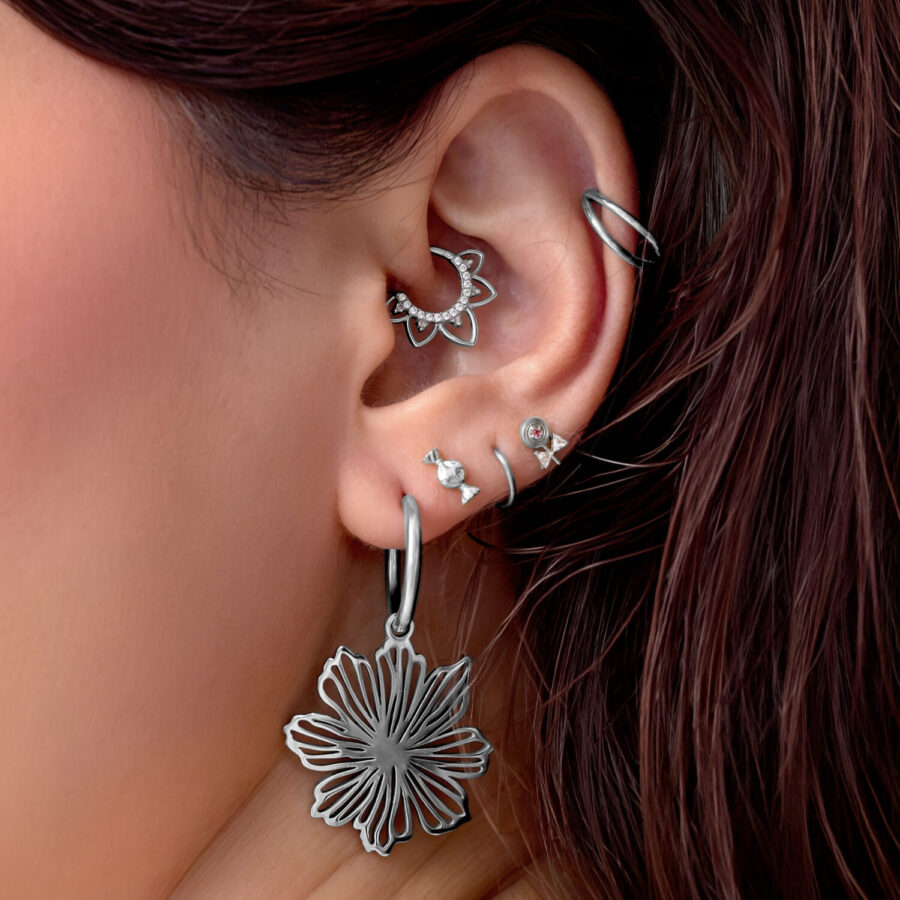 Primer plano de una oreja con varios piercings plateados, incluyendo un gran colgante de flor y un anillo delicado.