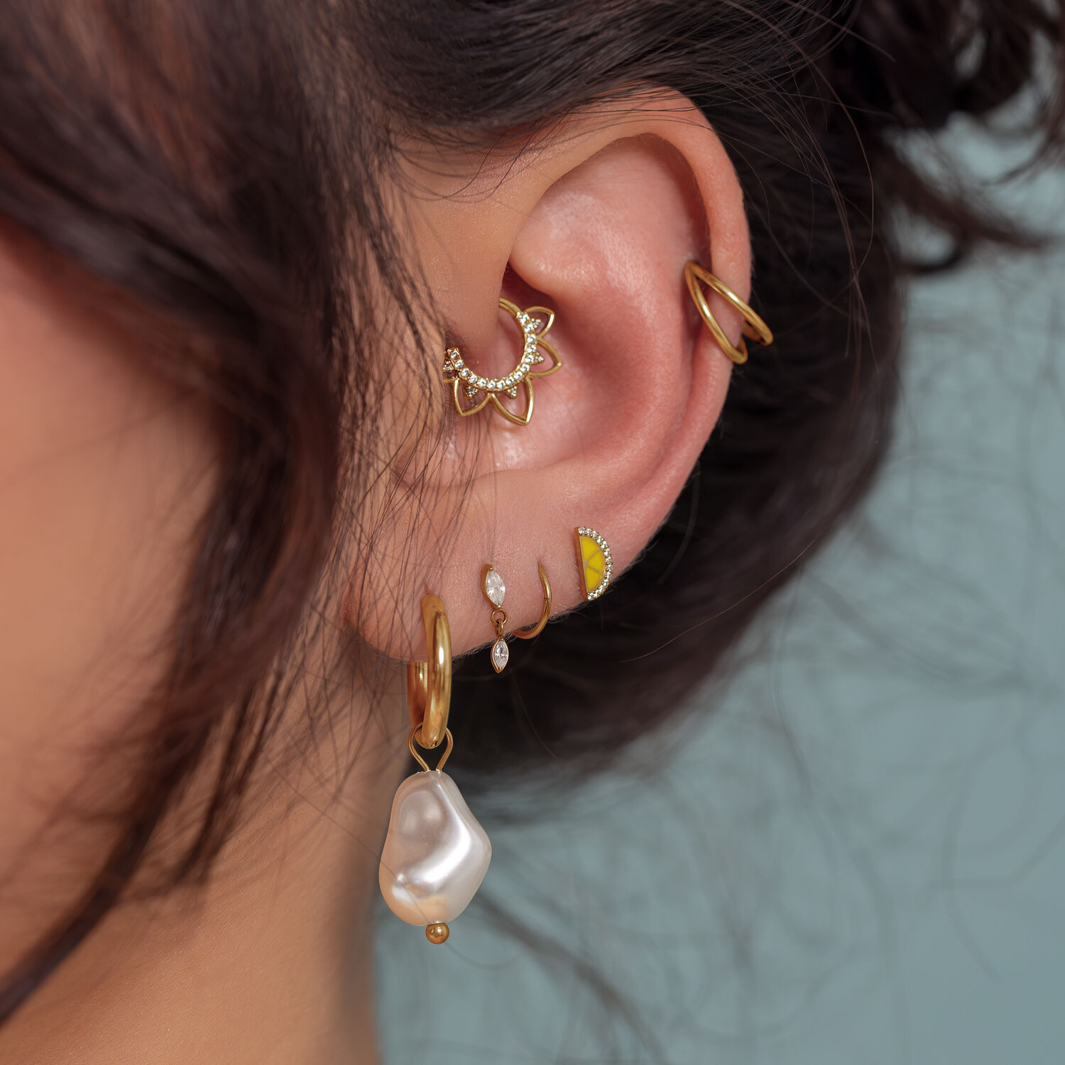 Piercings de oro para orejas con colgante de perla Primer plano de una oreja con varios piercings dorados, incluido un colgante de perla, sobre un fondo azul claro