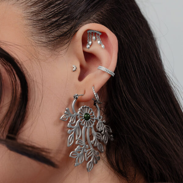 Primer plano de una oreja con varios piercings plateados, incluyendo un gran colgante ovalado y brillantes accesorios de botón.