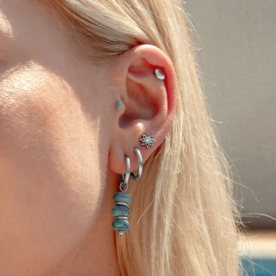 Primer plano de una oreja con pendientes de cristal turquesa y pequeños piercings plateados, piel de fondo