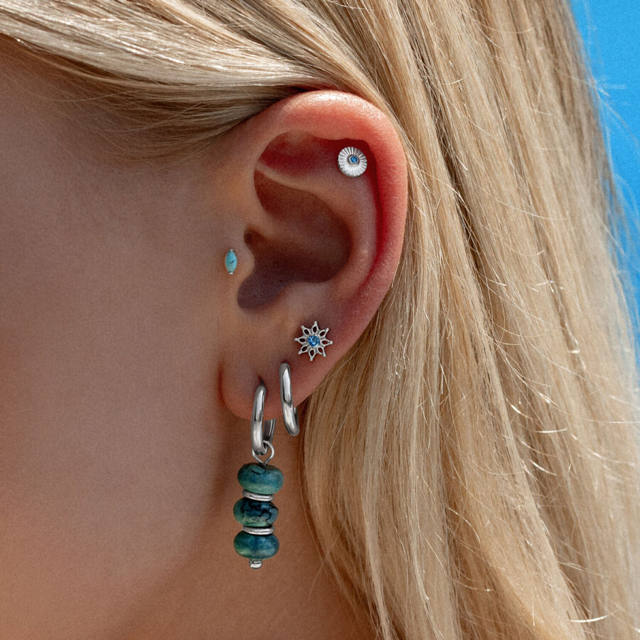 Oreja con varios piercings, incluyendo un ajuste de cristal turquesa y aros plateados, frente a un cielo azul