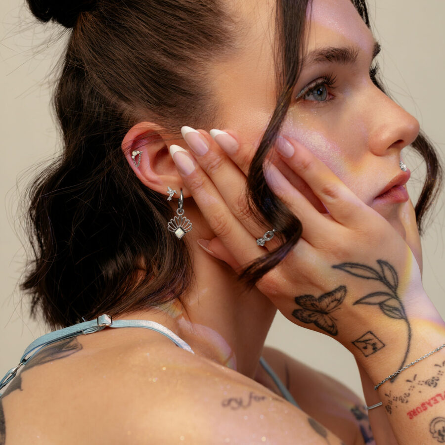 Joven con luz tenue en el rostro, lleva un pendiente de plata con colgante de mariposa y tiene tatuajes en las manos.