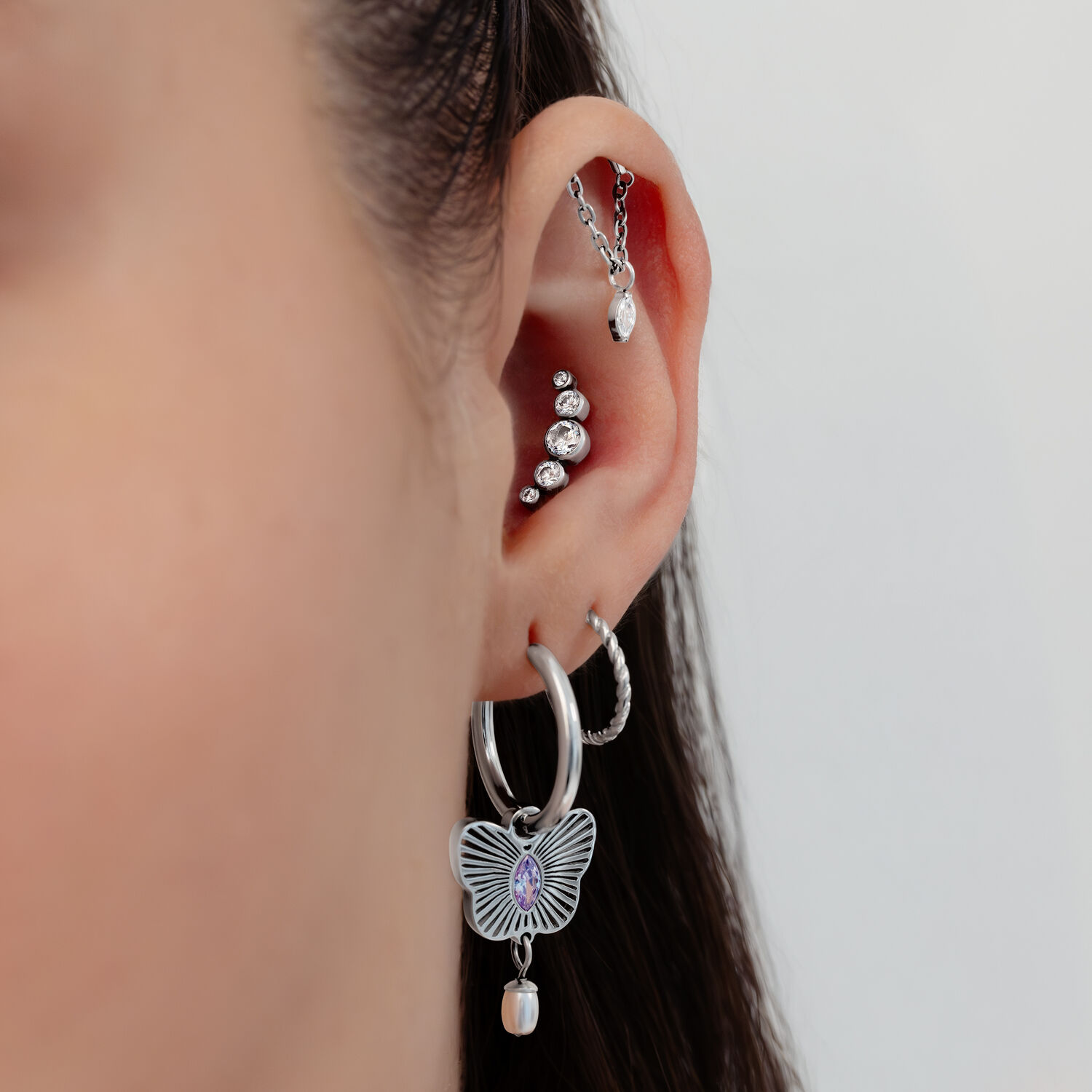 Piercings de oreja plateados con cristales Primer plano de una oreja con varios piercings plateados, cristales y un colgante de mariposa sobre un fondo blanco.