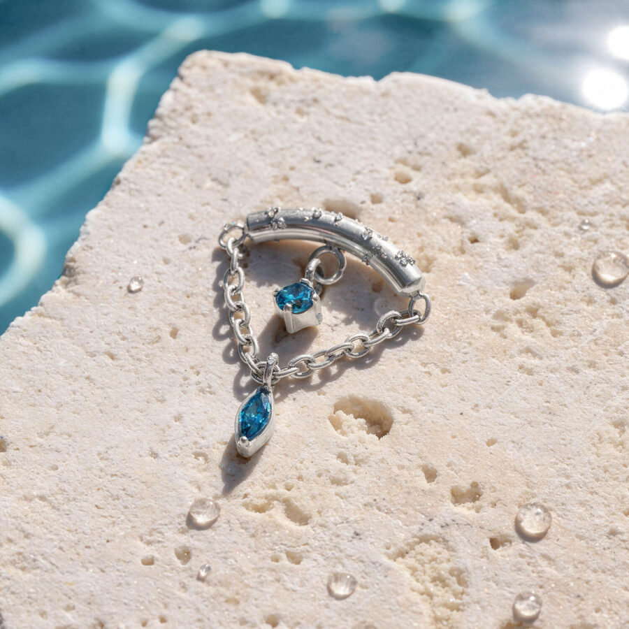 Adorno de piercing de hélice plateado con dos piedras turquesas sobre piedra clara con gotas de agua