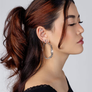 Mujer de cabello castaño lleva grandes aros de plata con diseño floral sobre un fondo gris.