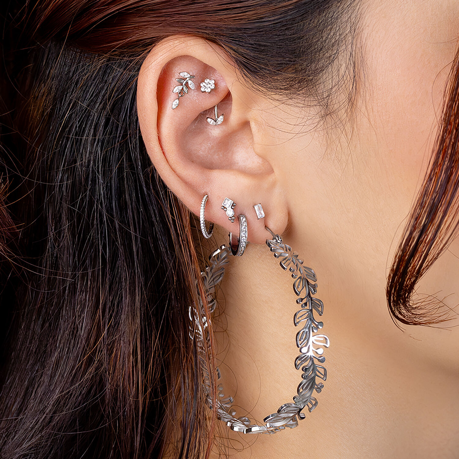 Pendientes de plata con cristales Oreja con varios piercings, incluyendo aros de plata y cristales, cabello castaño de fondo