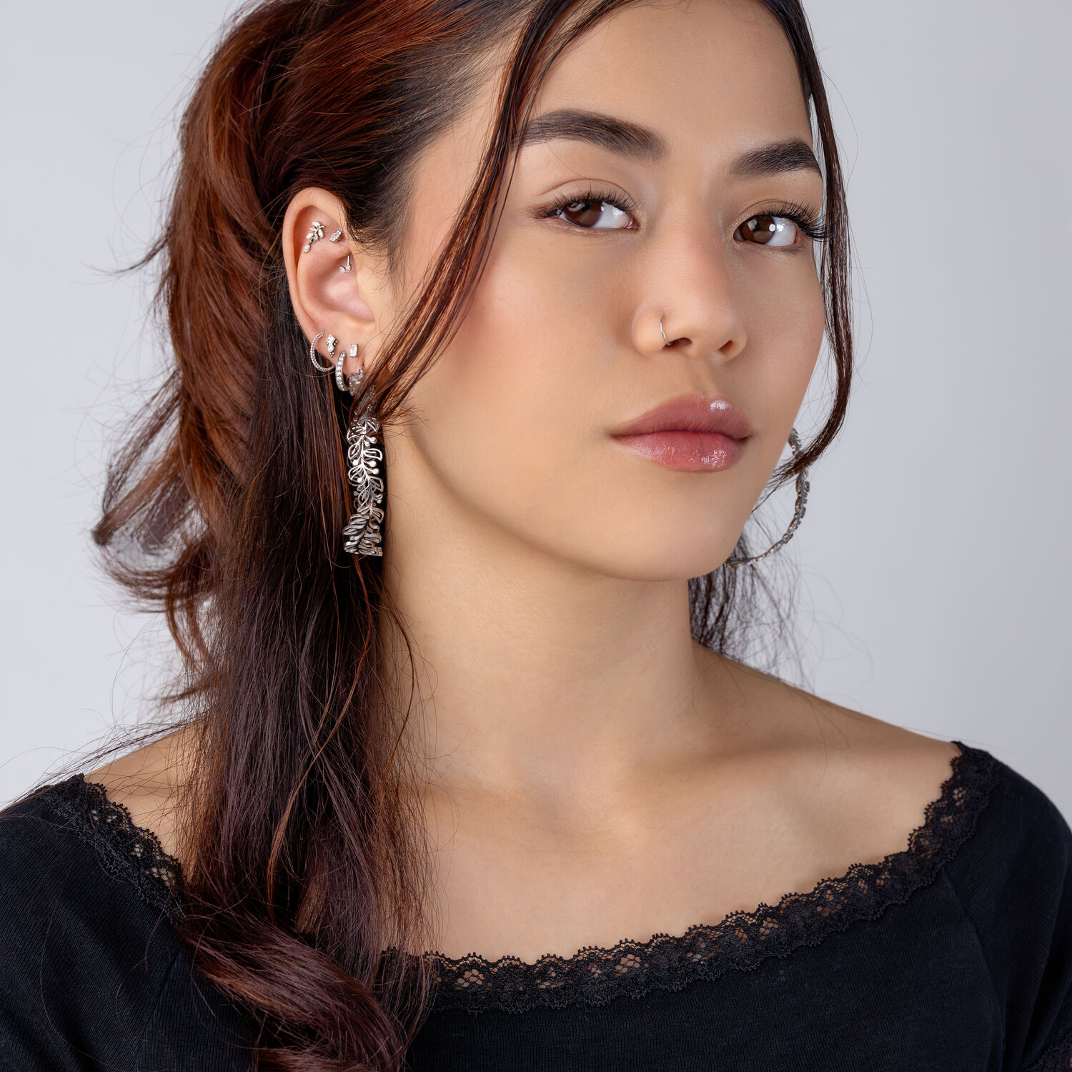 Piercings de oreja plateados con colgante Mujer con cabello largo y castaño lleva piercings de oreja plateados y suéter negro frente a un fondo gris