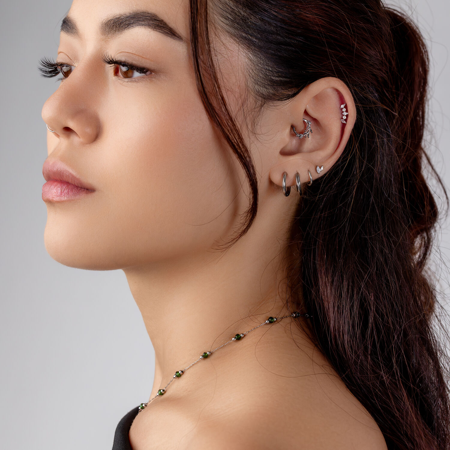 Piercing Helix de Plata con Cristales Mujer con varios piercings de plata en la oreja, incluyendo un Helix adornado con cristales, sobre un fondo gris.