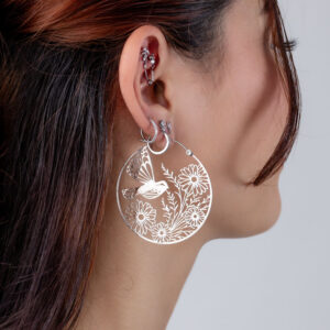 Pendientes plateados con diseño de flores y pájaros, llevados por una mujer con cabello castaño.