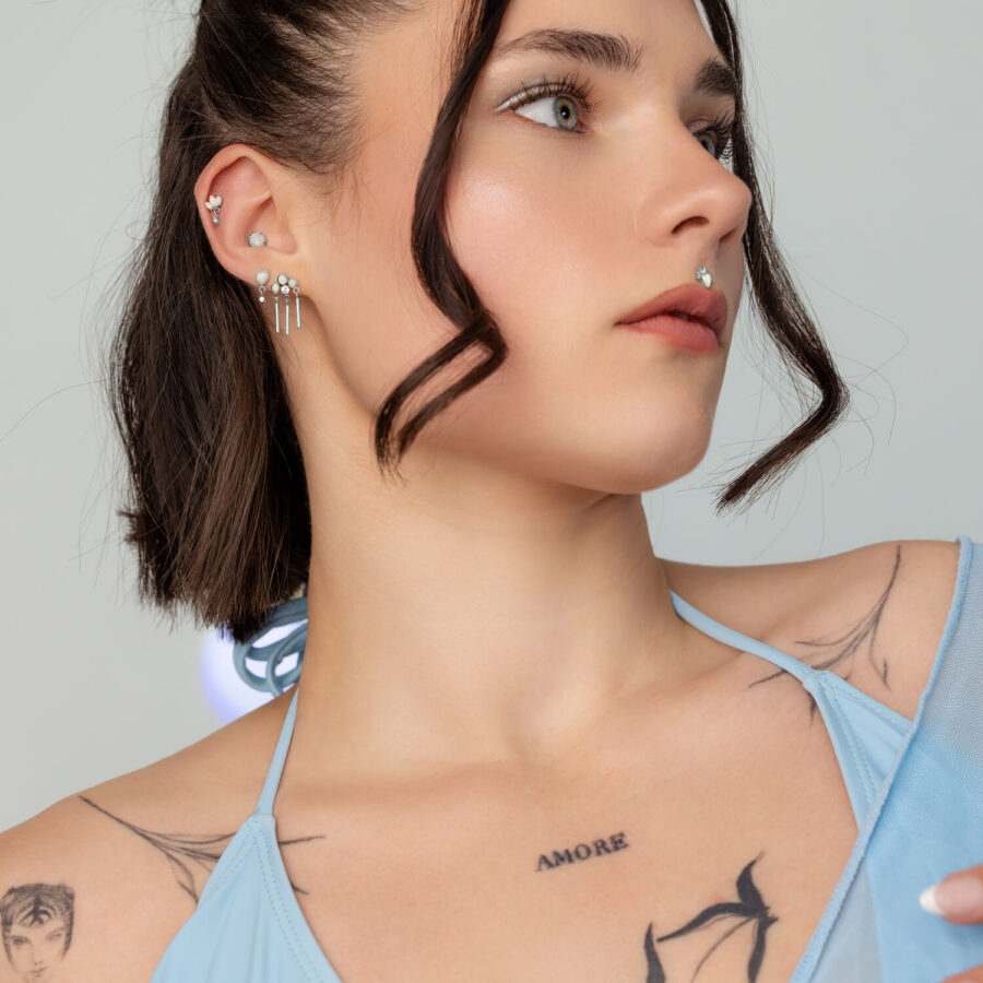 Mujer con varios accesorios de piercing plateados en la oreja, piedras de ópalo y blusa azul claro
