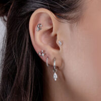 Primer plano de una oreja con varios piercings plateados y piedras cristalinas, sobre un fondo de cabello castaño.