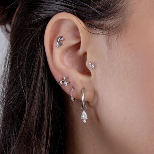 Primer plano de una oreja con varios piercings plateados y piedras cristalinas, sobre un fondo de cabello castaño.